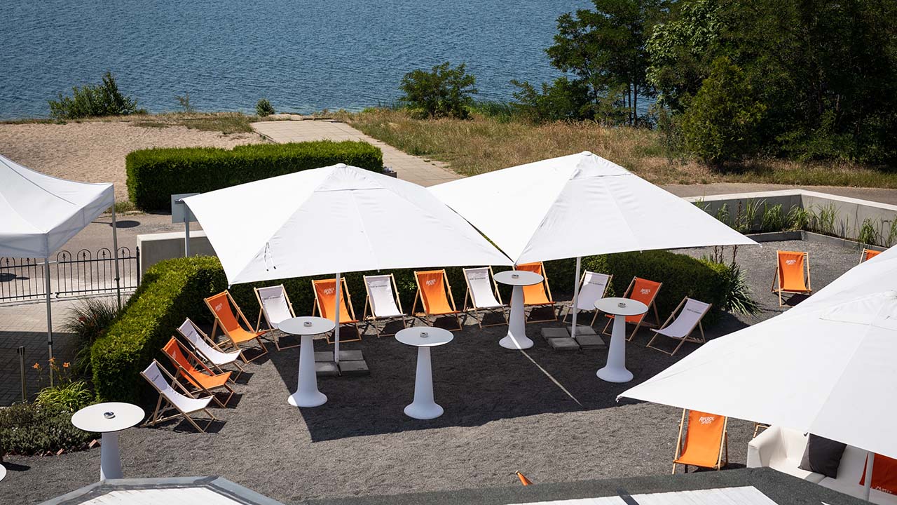 Als Teil unserer Bildergalerie zeigt diese Aufnahme den Außenbereich mit weißen und orangefarbenen Liegestühlen, weißen Stehtischen und Sonnenschirmen. Im Hintergrund ist das Seeufer zu sehen.