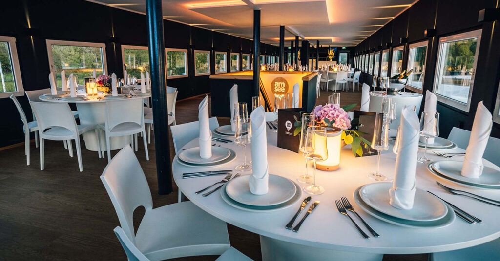Das Oberdeck der Eventlocation amSee ist eingerichtet mit weißen modernen Bankettmöbeln. Auf den Tischen stehen Blumenschmuck, Geschirr, Gläser sowie Stoffservietten gefaltet als Kerze.
