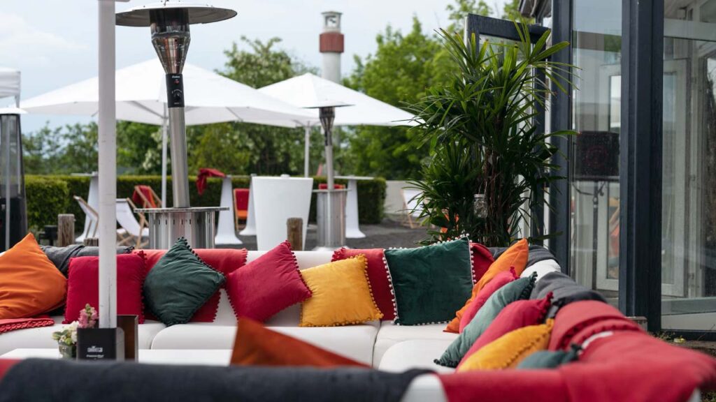 weiße Lounge-Sitzgruppe vor dem Wintergarten, bestückt mit Kissen und Decken in Rot, Orange und Anthrazit.