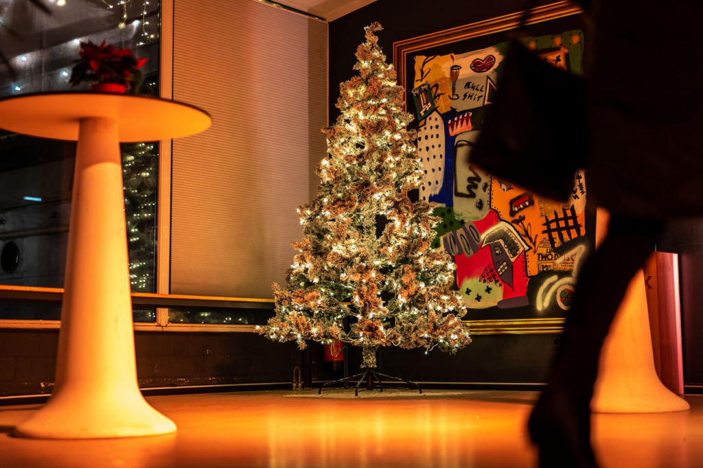Ein geschmückter Weihnachtsbaum sowie ein weißer runder Stehtisch im Wintergarten des Schiffs. Im Vordergrund läuft eine Person vorbei.