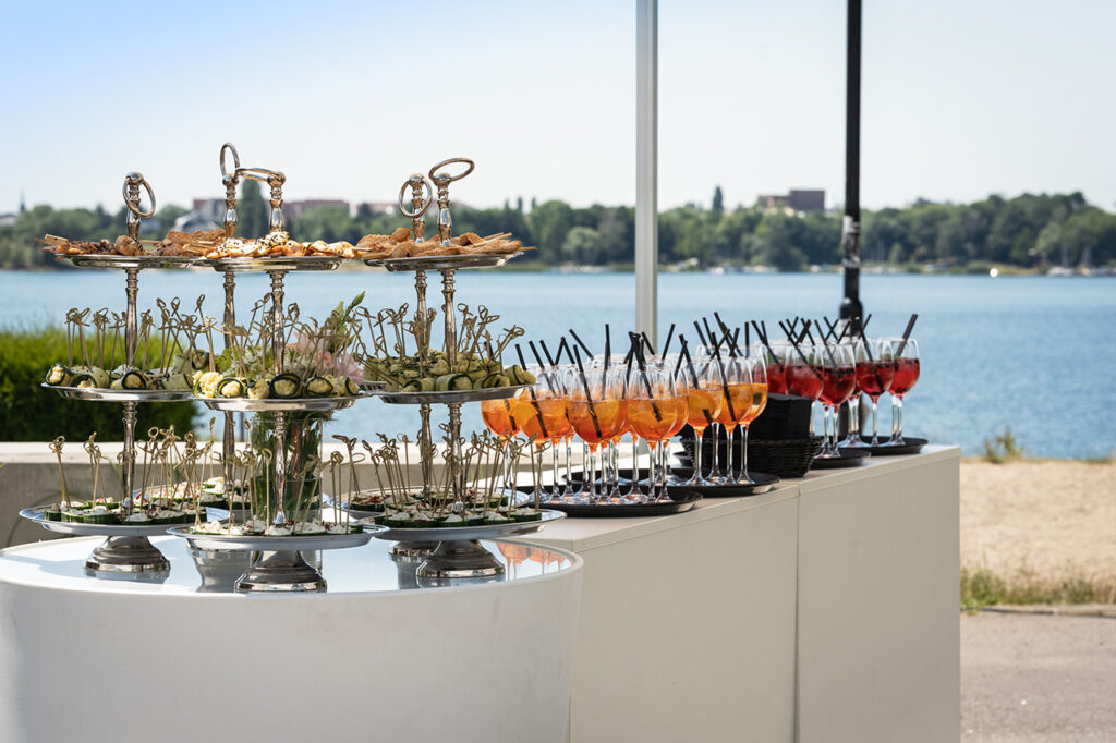 Empfangssituation an der amSee Location: weiße Counter mit Etageren mit Canapées sowie Gläsern mit Aperitif, im Hintergrund der See.
