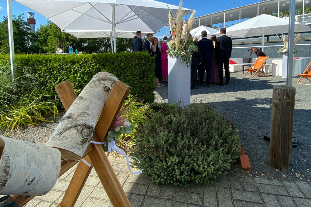 Hochzeitsfeier amSee, Im Hintergrund stellen sich die Gäste für ein Foto auf, im Vordergrund steht ein Baumstamm, der vom Paar zersägt wurde.