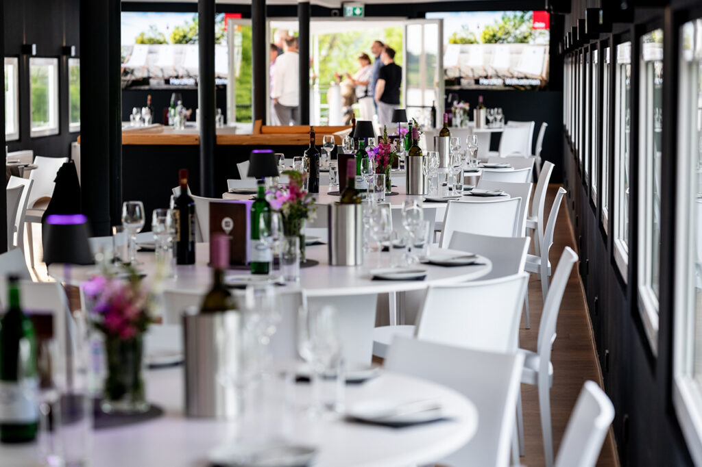 Das Oberdeck der Eventlocation amSee ist eingerichtet mit weißen modernen Bankettmöbeln. Auf den Tischen stehen Blumenschmuck, Geschirr, Gläser sowie Wein- und Wasserflaschen.