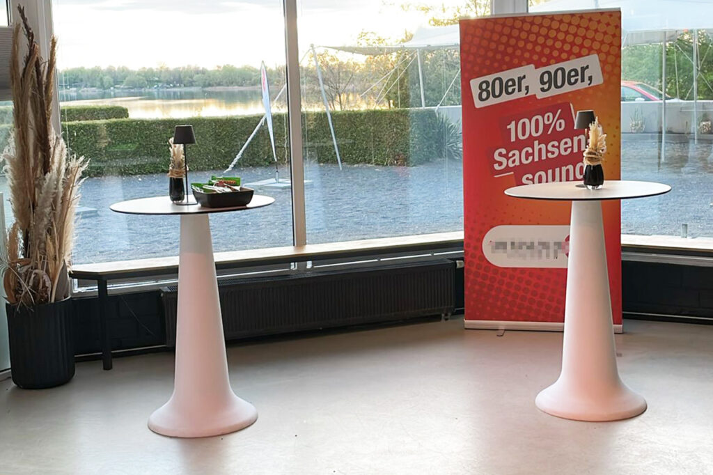 Produktpräsentation in der amSee Location Leipzig: Im Wintergarten mit Blick auf den See stehen Stehtische und ein Rollup eines Radiosenders.
