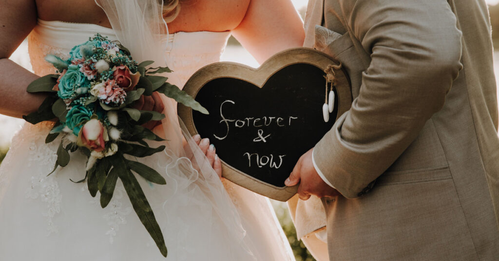 Ein Hochzeitspaar hält eine herzförmige Tafel mit der Aufschrift "Forever & now" in den Händen, daneben ein großen Blumenbouquet. Das Bild illustriert den Artikel über den Traumhochzeit-Ablauf in der Hochzeitslocation amSee Leipzig.