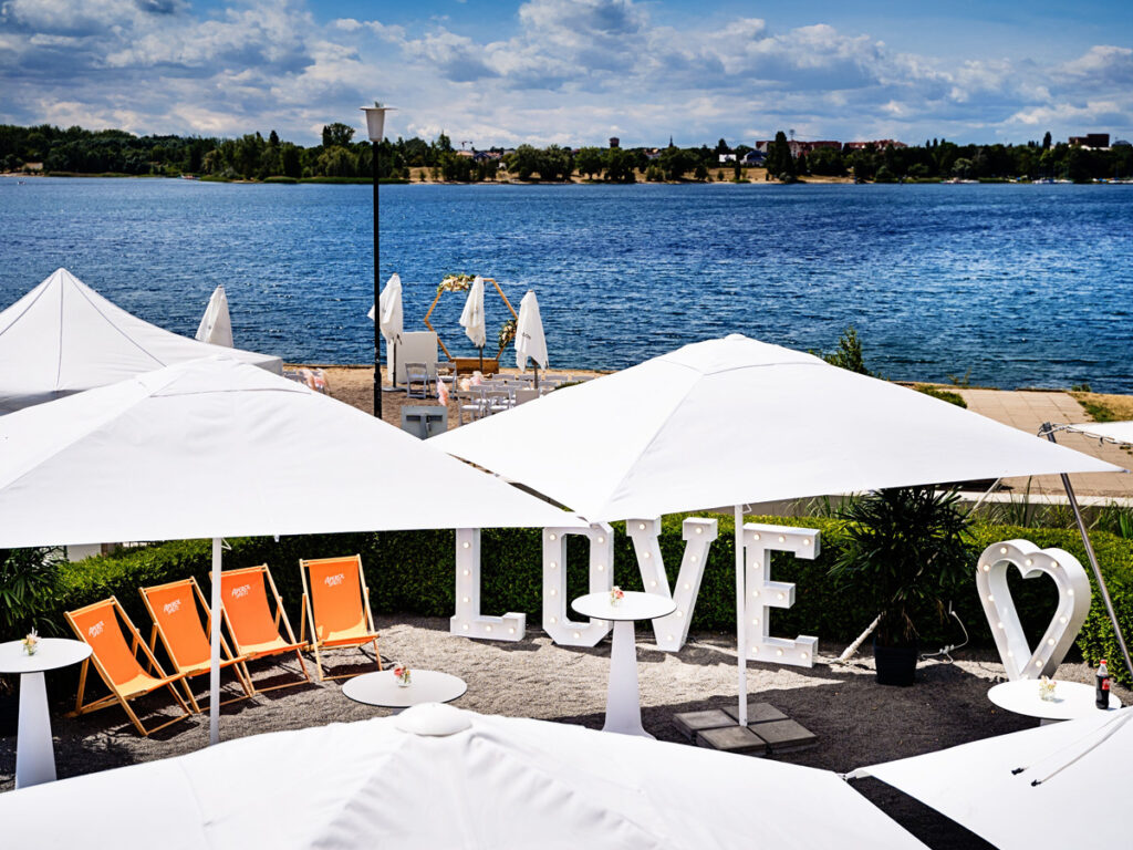 Traumhochzeit amSee: Empfangssituation bei strahlendem Sonnenschein, im Hintergrund der tiefblaue See und das Westufer, im Vordergrund weiße Schirme und weiße Leuchtbuchstaben "LOVE"