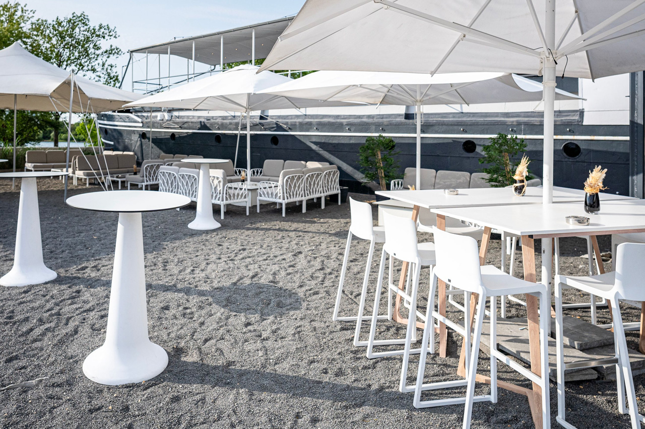 Seeterrasse der amSee Eventlocation Leipzig: weiße Stehtische, Barhocker, Lounges, im Hintergrund dunkelgrauer Schiffsrumpf