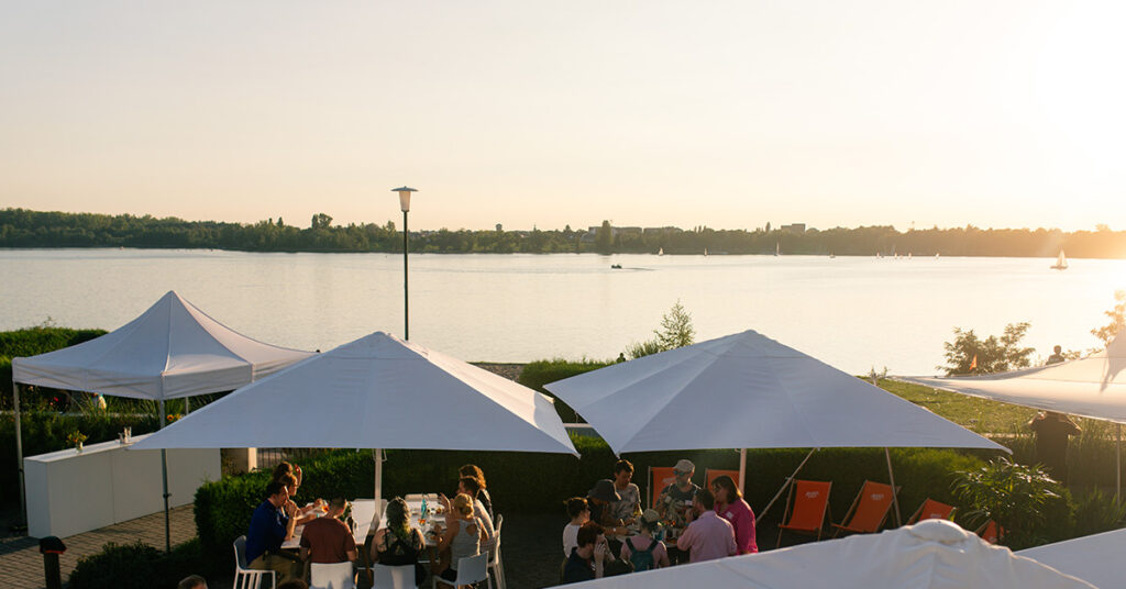 Eventlocation am See finden Leipzig: Blick von der Location amSee über den Außenbereich mit weißen Schirmen und Eventgästen in Richtung See