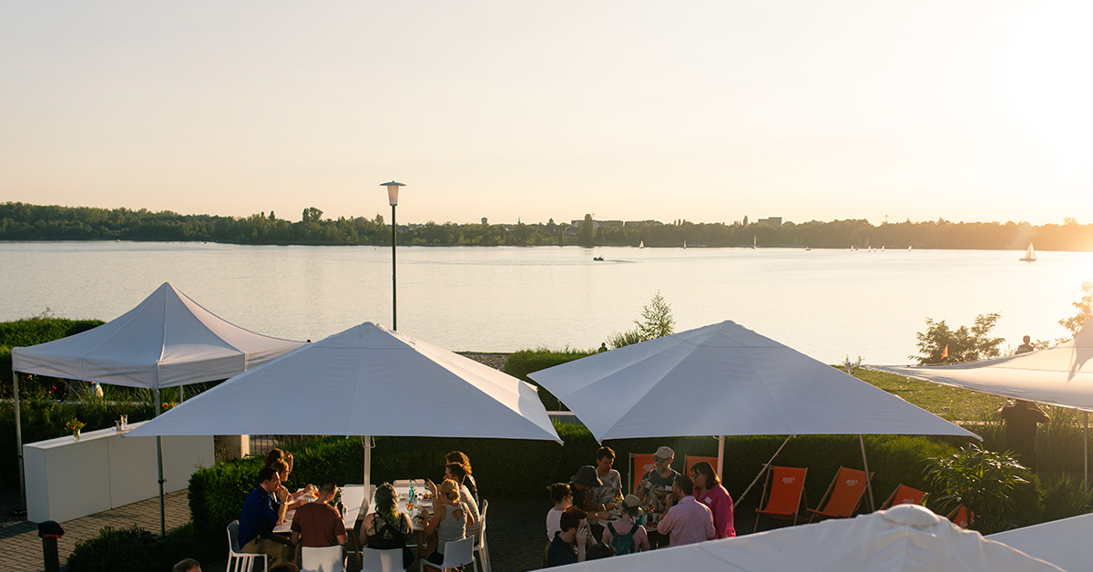 Eventlocation am See finden Leipzig: Blick von der Location amSee über den Außenbereich mit weißen Schirmen und Eventgästen in Richtung See